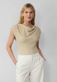 Beige textuur top met een gedrapeerde halslijn en korte mouwen, gecombineerd met witte hoge taille broeken. Heeft een relaxte pasvorm en een subtiel patroon.