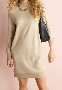 Robe sweatshirt beige avec des manches longues, col et ourlet côtelés, équipée d'une fermeture éclair sur le côté, associée à un sac à épaule noir et un collier en chaîne.
