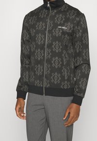 Chaqueta negra con cremallera y un elaborado patrón dorado, cuello alto y puños acanalados. Presenta el logo "LES DEUX" en el pecho. Textura de tela suave.