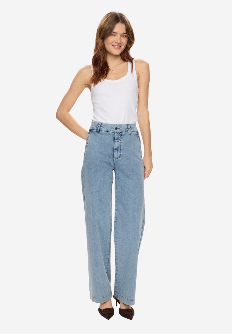 Jeune femme aux longs cheveux bruns portant un débardeur blanc, un jean large bleu clair taille haute et des escarpins à bout pointu marron foncé, souriante.