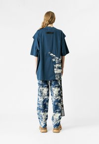 Persona vistiendo una camisa azul holgada con un parche abstracto blanco y azul y pantalones a juego, de pie frente a un fondo blanco liso.