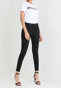 Kvinna iklädd en vit "Miss Sixty" T-shirt, svarta skinny jeans och svarta högklackade sandaler, stående mot en enfärgad vit bakgrund.