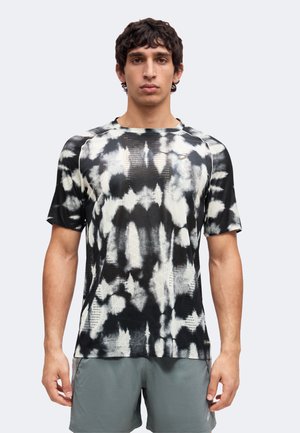 Ung mand stående forfra iført en sort og hvid tie-dye kortærmet skjorte og grå træningsshorts mod en ensfarvet baggrund.