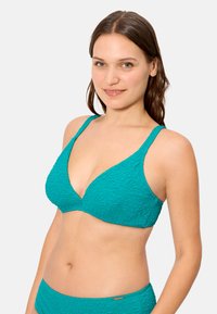 Mujer joven con cabello castaño que lleva un bikini texturizado color verde azulado, con la parte superior y el pantalón a juego, sonriendo, sobre un fondo claro y sencillo.