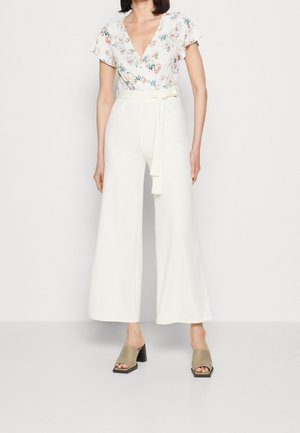Kvinde iført hvide bukser med brede ben og bindebånd i taljen, blomsterprintet kortærmet V-hals bluse og beige blokhælede sandal med åben tå, stående mod en hvid baggrund.