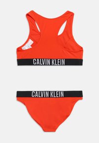 Φωτεινό πορτοκαλί αθλητικό σουτιέν Calvin Klein και ταιριαστό σλιπ με μαύρες ελαστικές ταινίες που φέρουν λευκό λογότυπο της μάρκας, απλωμένα επίπεδα σε λευκό φόντο.