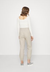 Femme aux cheveux bruns mi-longs portant un haut blanc à manches longues et un pantalon beige taille haute avec des poignets élastiques, debout de dos.