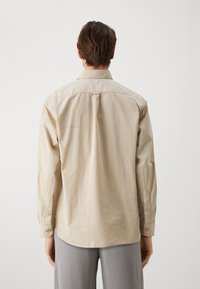 Camisa de manga larga beige con corte recto, puños abotonados y un pliegue en la espalda. Fabricada con una tela suave y ligera.