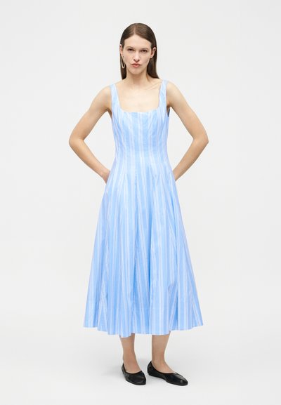 Γυναίκα που φοράει αμάνικο ανοιχτό μπλε φόρεμα με λευκές κάθετες ρίγες, μαύρα ίσια παπούτσια και χρυσά γεωμετρικά σκουλαρίκια, στέκεται με τα χέρια στην πλάτη.