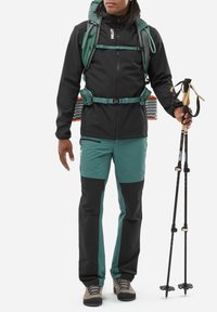 Veste en tissu soft-shell noire avec des accents teal, associée à un pantalon teal et noir. Équipée d'un sac à dos vert et de bâtons de trekking.
