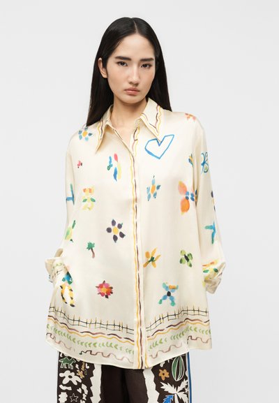 Νεαρή γυναίκα με μακριά σκούρα μαλλιά που φοράει ένα άνετο κρεμ πουκάμισο με πολύχρωμα αφηρημένα ανθικά και καρδιές μοτίβα σε απλό φόντο.