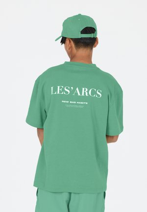Mintgrön oversized t-shirt med korta ärmar, med vit text "LES'ARCS" och "NEW BAD HABITS" på baksidan. Mjuk bomullsmaterial.