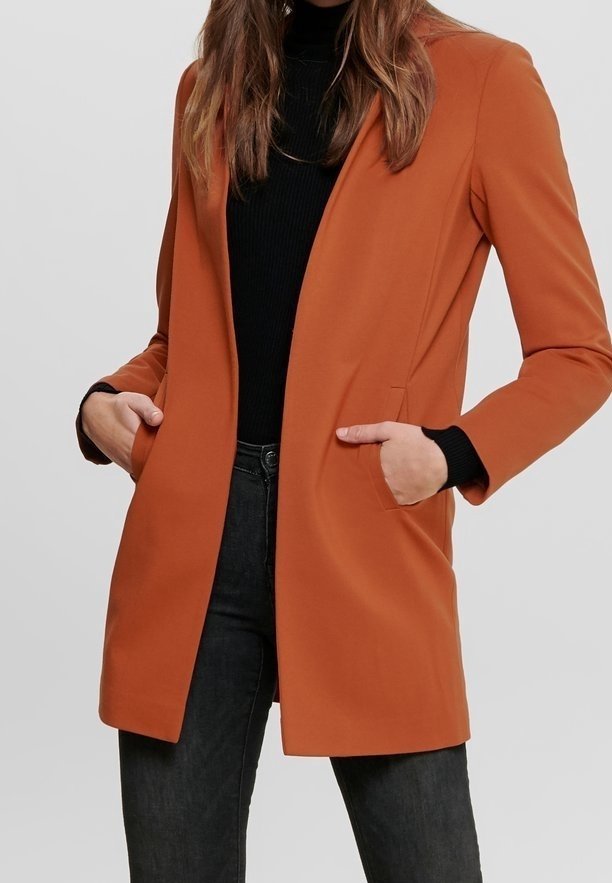 Femme portant un long blazer orange brûlé, les mains dans les poches, associé à un col roulé noir et un jean foncé.