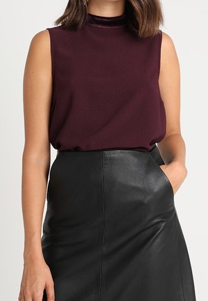 Top sin mangas de color púrpura oscuro con cuello alto, combinado con una falda de cuero negra, con una mano en el bolsillo de la falda.