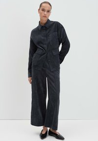 Donna in giacca di velluto a coste grigia scura con grandi tasche e pantaloni a gamba larga abbinati, in piedi contro uno sfondo bianco semplice.