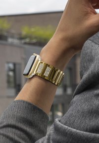 Montre en métal doré avec un écran noir brillant, mettant en avant des accents scintillants sur le bracelet. Le bracelet a une finition brillante et maillée.