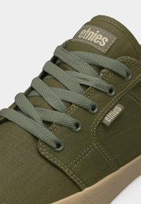 Baskets Etnies vert olive avec lacets plats, dessus en tissu, détails de couture, et semelle en caoutchouc beige.