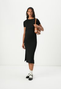 Μαύρο ribbed midi φόρεμα με κοντά μανίκια και πλευρικό σκίσιμο, συνδυασμένο με καφέ τσάντα. Το μοντέλο φοράει λευκές κάλτσες και μαύρα παπούτσια.