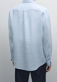 Camicia a maniche lunghe blu chiaro in tessuto leggero con yoke posteriore, orlo arrotondato e polsini con bottoni. Texture liscia, design minimalista.