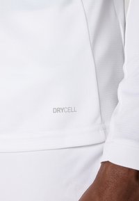 Hvit atletisk topp laget av teksturert stoff. Har "DRYCELL"-logo i grått. Lange ermer med avslappet passform. Enkel, funksjonell design.