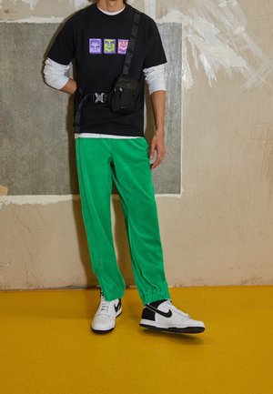 Czarny t-shirt z kolorowymi nadrukami graficznymi, zielone dresowe spodnie z weluru, białe sneakersy z czarnymi akcentami i mała czarna torba na ramię.