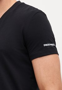 Schwarzes kurzärmliges T-Shirt mit V-Ausschnitt, aus glattem Stoff gefertigt. Mit einem weißen Logo am Ärmel, im minimalistischen Stil.