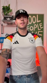 Man wearing Germany soccer jersey and black cap standing indoors with plant, shoes, and green sign in the background.