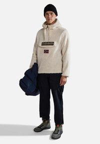 Felpa oversized color crema in pile con zip, dotata di tasca frontale e toppe; berretto nero; pantaloni cargo; giacca imbottita in mano; stivali verdi e grigi.