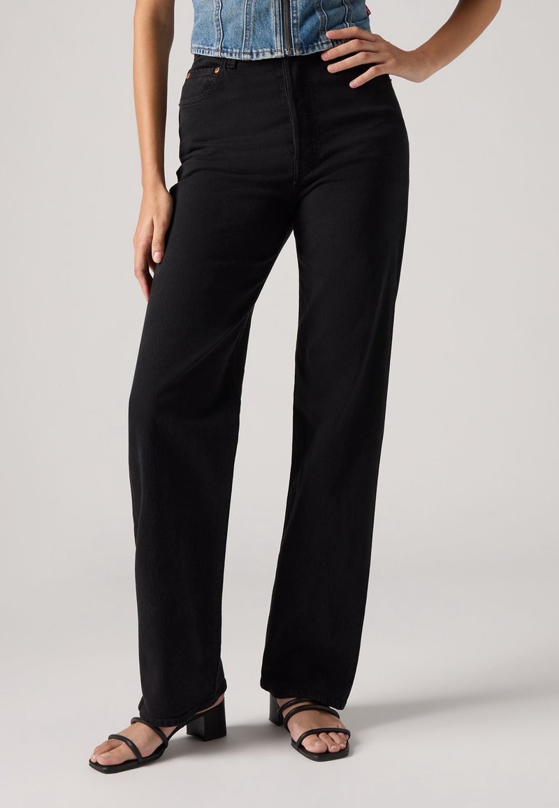 Jean noir taille haute à jambes larges en denim, avec une fermeture éclair à l'avant, des passants de ceinture et des coutures minimales. Associé à des sandales noires à lanières.