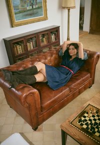 Moteris mėlynoje suknelėje ir juodais batais atsilošusi ant rudos odinės sofos kambaryje su medine knygų lenta ir šachmatų lenta ant stalo.