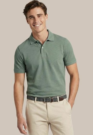 Lachende jonge man met een groen korte mouw polo, beige broek en een zwart-witte geweven riem, staand met één hand in de zak.