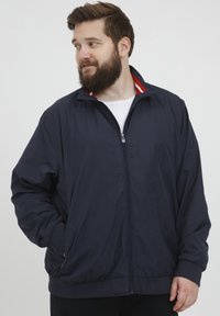 Jachetă de sport de culoare bleumarin, confectionată dintr-un material ușor, având închidere cu fermoar, manșete ribbate și un guler cu dungi roșii. Include două buzunare laterale.