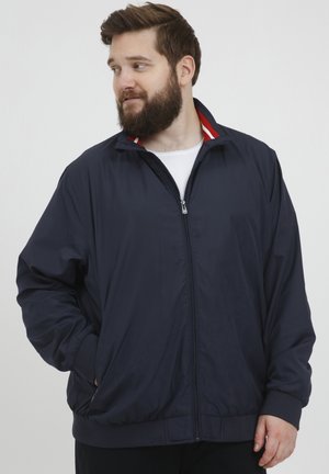 Jachetă de sport de culoare bleumarin, confectionată dintr-un material ușor, având închidere cu fermoar, manșete ribbate și un guler cu dungi roșii. Include două buzunare laterale.