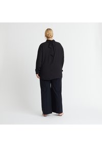 Schwarze Bluse mit langen Ärmeln und einer gebundenen Schleife am Hals, kombiniert mit weit geschnittenen schwarzen Hosen. Glatter Stoff und lockere Passform.