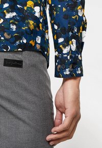 Flerfärgad skjorta med blå, gul, brun och vit abstrakt mönster; har en knappfäste vid ärmslutet. Gråa tygbyxor med en strukturerad yta.