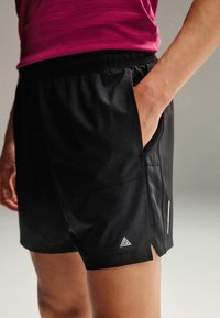 Personne portant un short de sport noir avec la main dans la poche et un t-shirt magenta visible de la taille vers le bas sur un fond neutre.