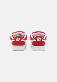 Baskets en suede rouge avec des accents blancs, dotées d'une semelle blanche texturée et d'un logo Puma doré près du talon. Design classique, style basse.