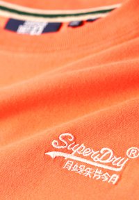 T-shirt in cotone arancione con tessuto testurizzato, logo "Superdry" ricamato in bianco e colletto a righe con accenti verdi e rossi.