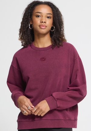 Ung kvinde med krøllet hår iført sølv creoløreringe og en bordeaux langærmet sweatshirt, der holder hænderne sammen i taljehøjde.