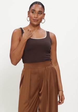 Woman with hoop earrings wearing a sleeveless dark brown top with light trim and high-waisted brown pleated pants against a plain background.