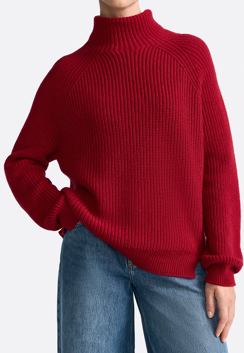 Rød strikket sweater med høj krave, ribbet tekstur og afslappet pasform. Lange ærmer og manchetter komplementerer designet. Bæres med blå jeans.