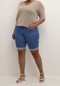 Pantalones cortos de mezclilla con puños enrollados, color azul claro y diseño de cinco bolsillos. Combinados con un top beige brillante de manga corta y sandalias con tiras.