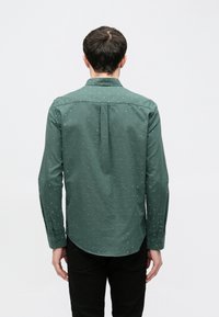 Grünes Langarmhemd mit Button-Down-Kragen, versehen mit einem Tupfenmuster in Hellblau und abgerundetem Saum mit seitlichen Schlitzen.