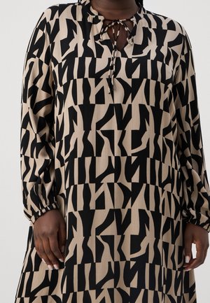 Personne portant une robe beige ample avec un imprimé géométrique abstrait noir audacieux, des manches longues et un col à nouer, debout devant un fond uni.