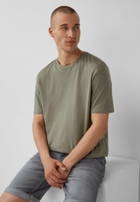 Olijfgroene t-shirt met korte mouwen en ronde hals, voorzien van een subtiele QS-logo. Model draagt lichte grijze denim shorts en zit op een witte pedestal.