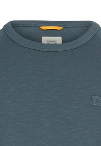 Czarna koszulka z ciemnoniebieskiego materiału o teksturowanym wykończeniu. Posiada prążkowany okrągły dekolt oraz małą pomarańczową metkę akcentującą przy kołnierzu. Na piersi wyszyte logo płaszcza.