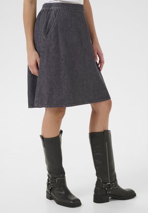 Vrouw draagt een donkergrijze rok tot op de knie met zijzakken en zwarte leren laarzen tot halverwege de kuit met riemen en gespen.