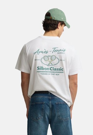 Joven con una camiseta blanca con raquetas de tenis y el texto "Après-Tennis Monaco Silbon Classic Tennis by the Sea" en la espalda, vaqueros azules y gorra verde.
