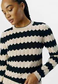 Strickpullover mit einem schwarz-weißen Zickzackmuster, gerippter Textur, rundem Ausschnitt und langen Ärmeln, die kontrastierende Bündchen aufweisen.