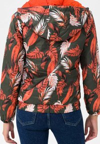 Huppari, jossa on tummanvihreä tausta ja rohkeat punaiset ja valkoiset lehtikuvioinnit. Varustettu joustavilla resoreilla hihoissa ja kerätyllä helmareunalla.